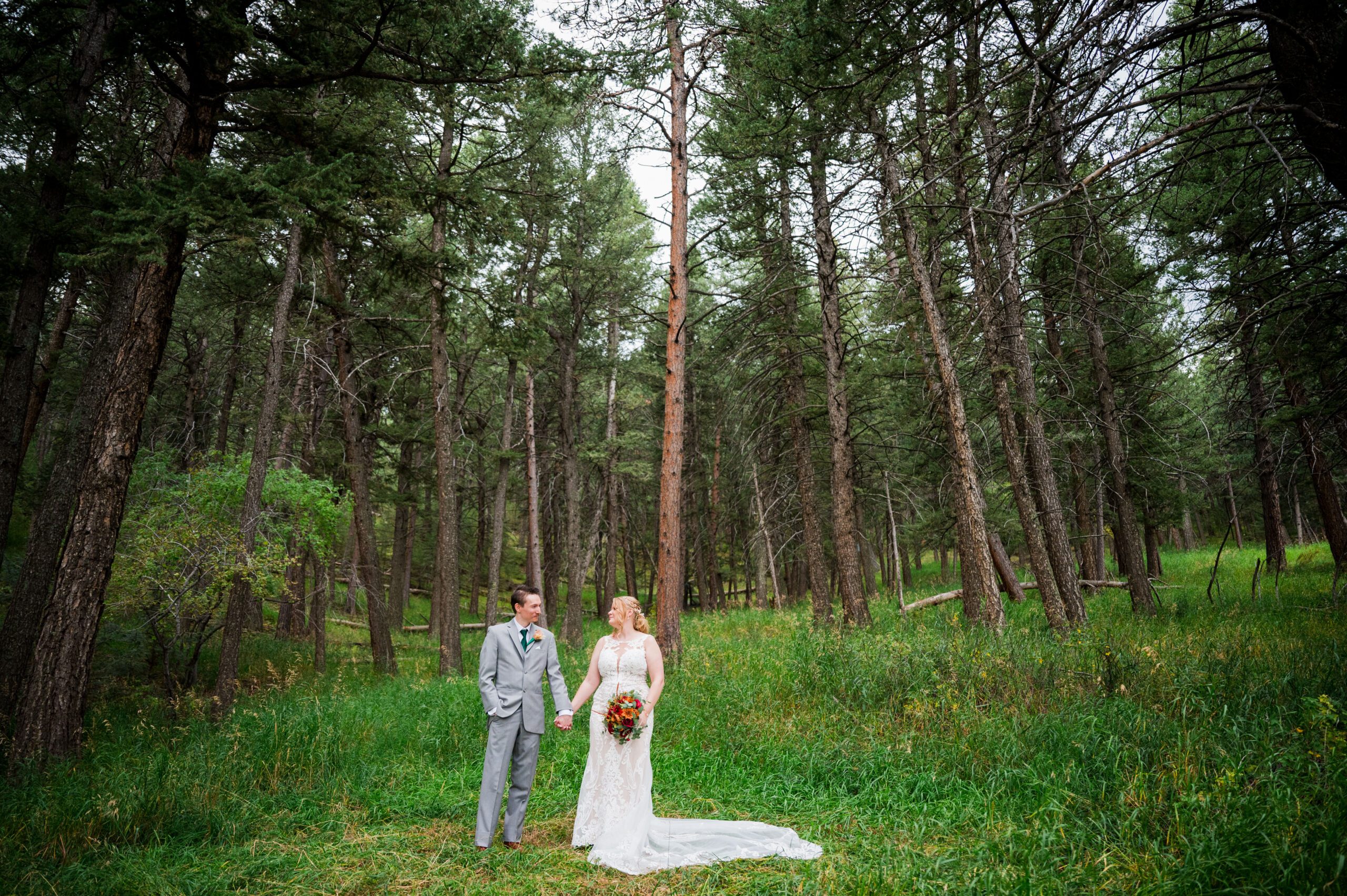 A bride and groom stand hand in hand looking at each other standing in a pine forest.